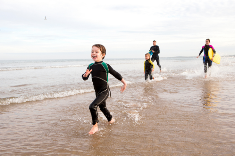 A family in wetsuits with surfboards at the beach. Family Friendly Motorhome and Caravan Holiday Ideas by Webbs.